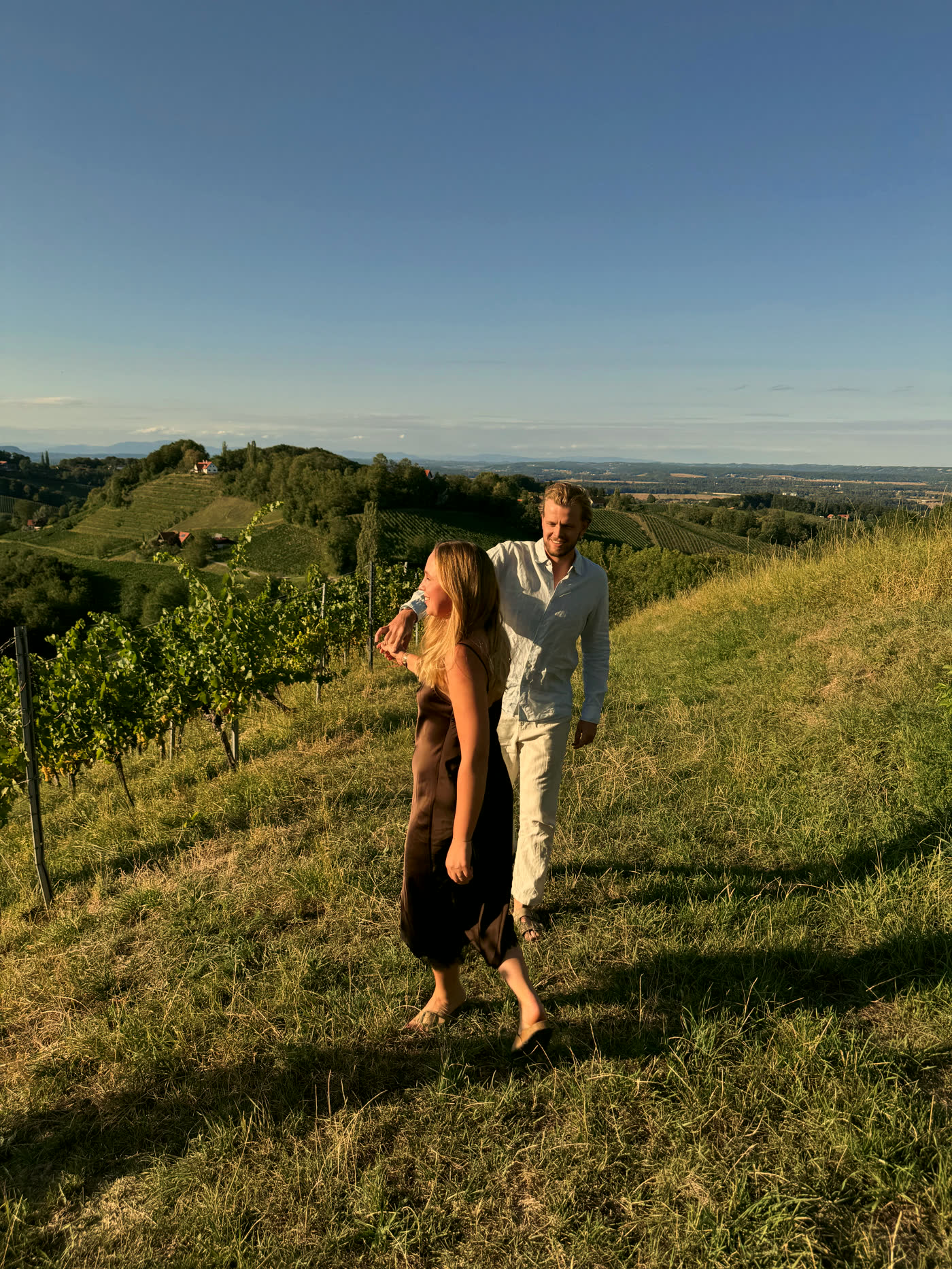 Ines und Oliver spazieren durch die Weingärten, dahinter der Blick über die Südsteiermark